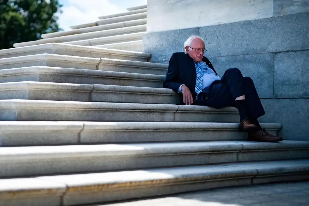 An older man with white hair and glasses, wearing a dark suit, light blue shirt, and patterned tie, sits slumped on wide…