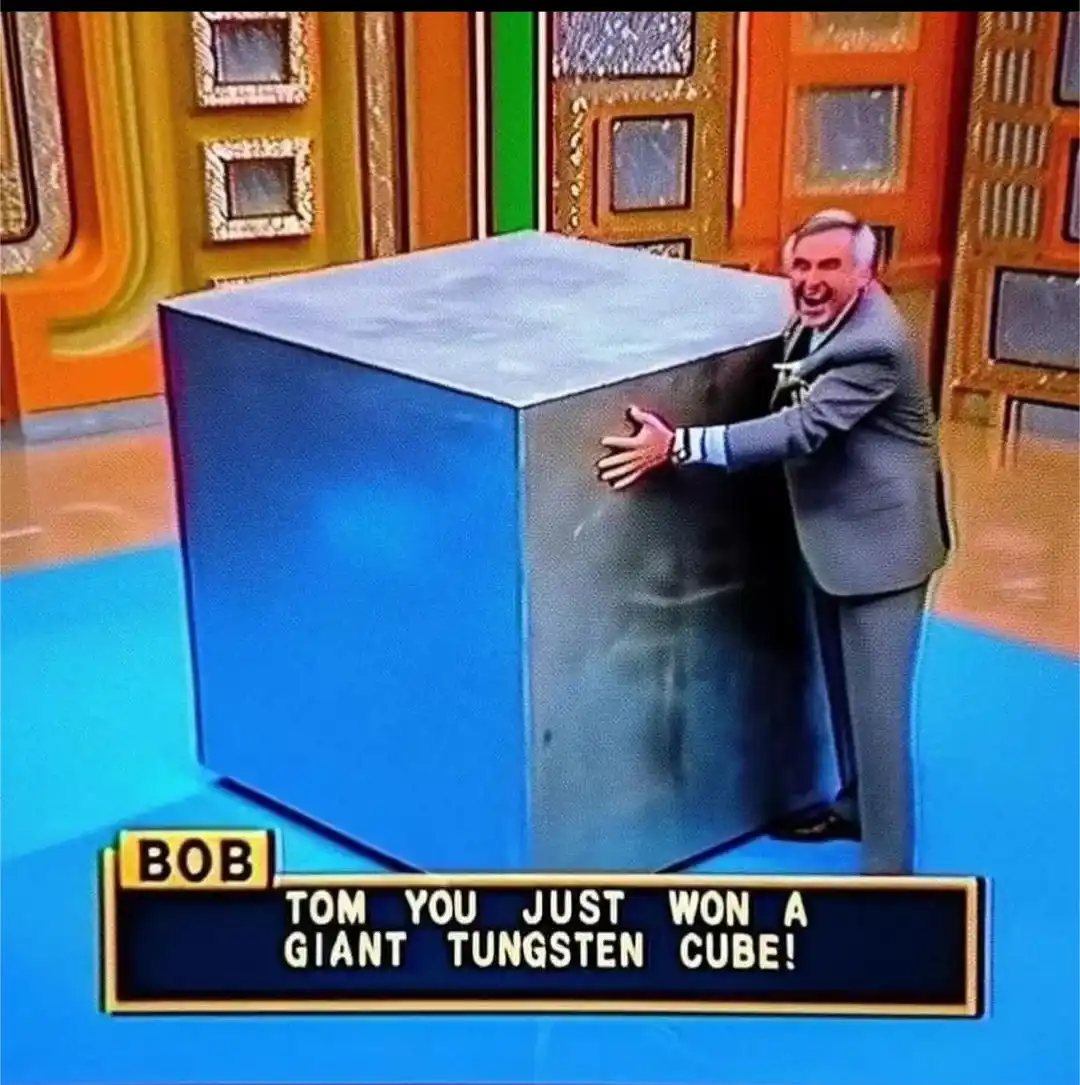 A man in a suit is shown struggling to push a giant, heavy-looking blue cube on a brightly colored game show set. A lower…