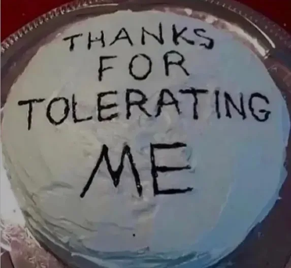 A close-up of a round cake with white frosting, sitting on a silver platter. The cake has the words 'THANKS FOR TOLERATING…