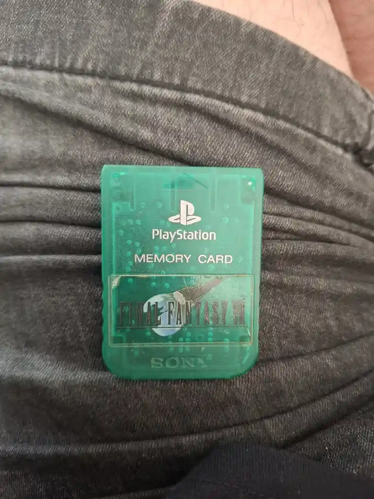 A translucent green PlayStation memory card for Final Fantasy VII is resting on a person's gray fabric clothing, showing the…