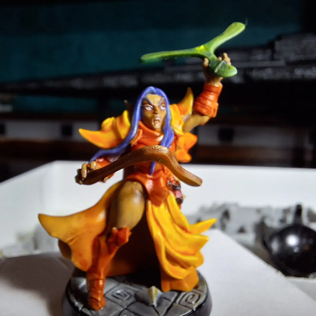 A painted fantasy mini-figure, resembling a warrior with blue hair, orange and yellow robes, and holding a green leaf-like…