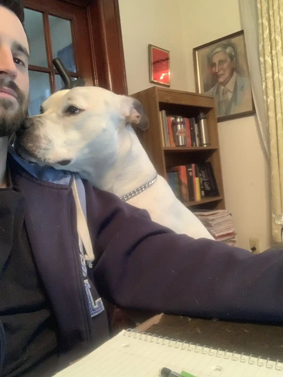A man with a beard takes a selfie while a white dog with a silver chain collar rests its head on his shoulder. In the…