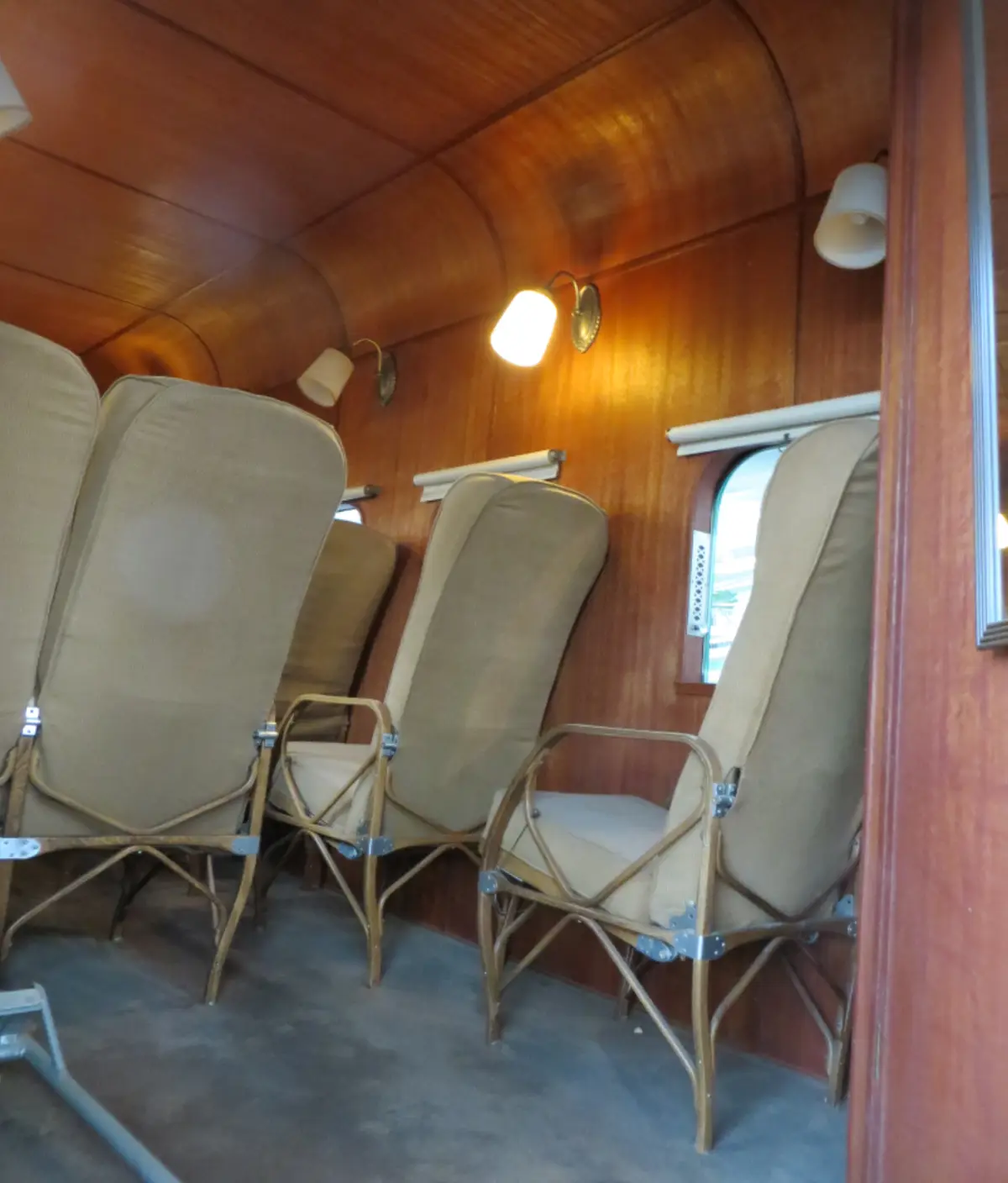 the image shows the interior of a vintage train car with wooden paneling, arched ceiling, and several beige upholstered…