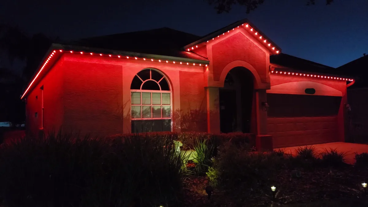 A single-story house at night is bathed in intense red light from string lights along its roofline and eaves, making the…