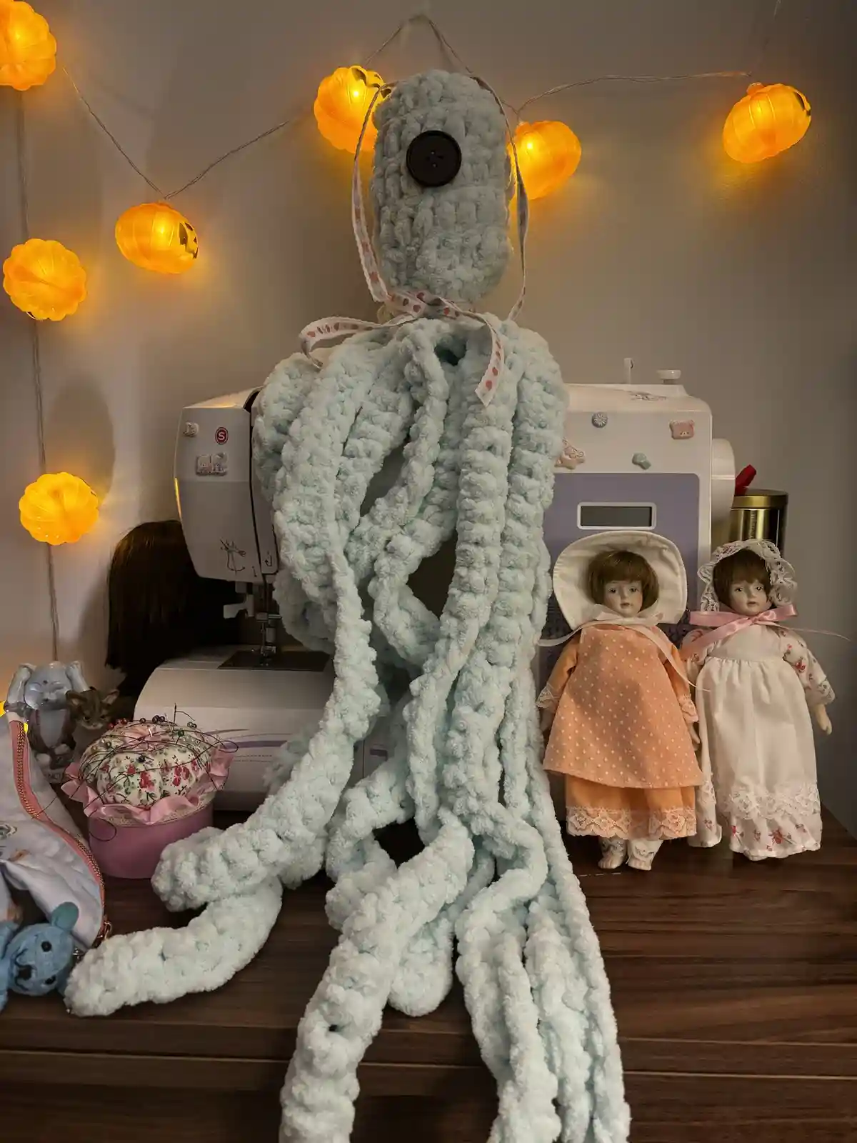 A large, light blue, fuzzy crocheted octopus-like creature with a black button eye sits on a wooden table in a craft room…