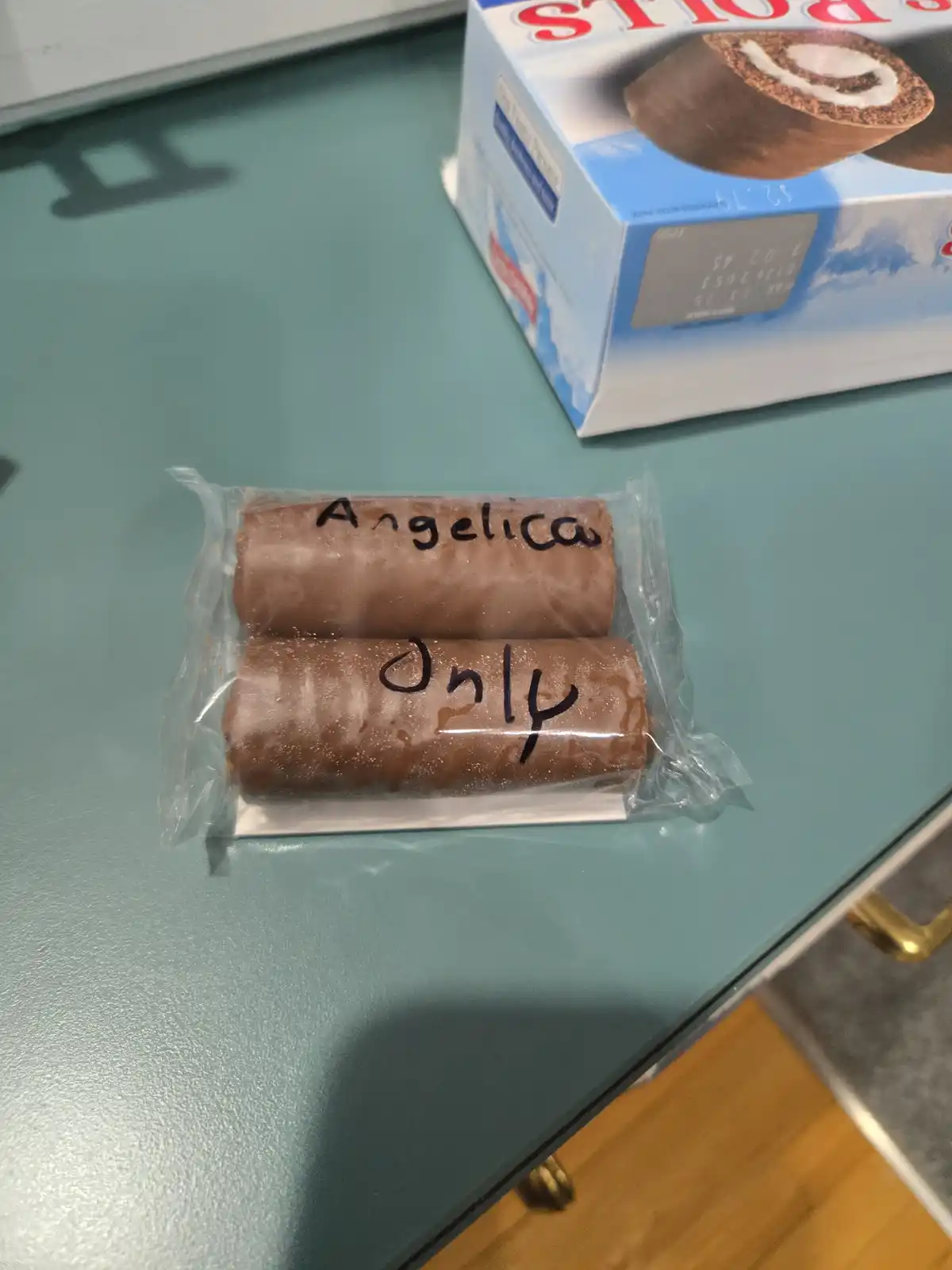 Two chocolate Swiss rolls in a clear plastic wrapper sit on a teal countertop, with the words 'Angelica Only' handwritten in…