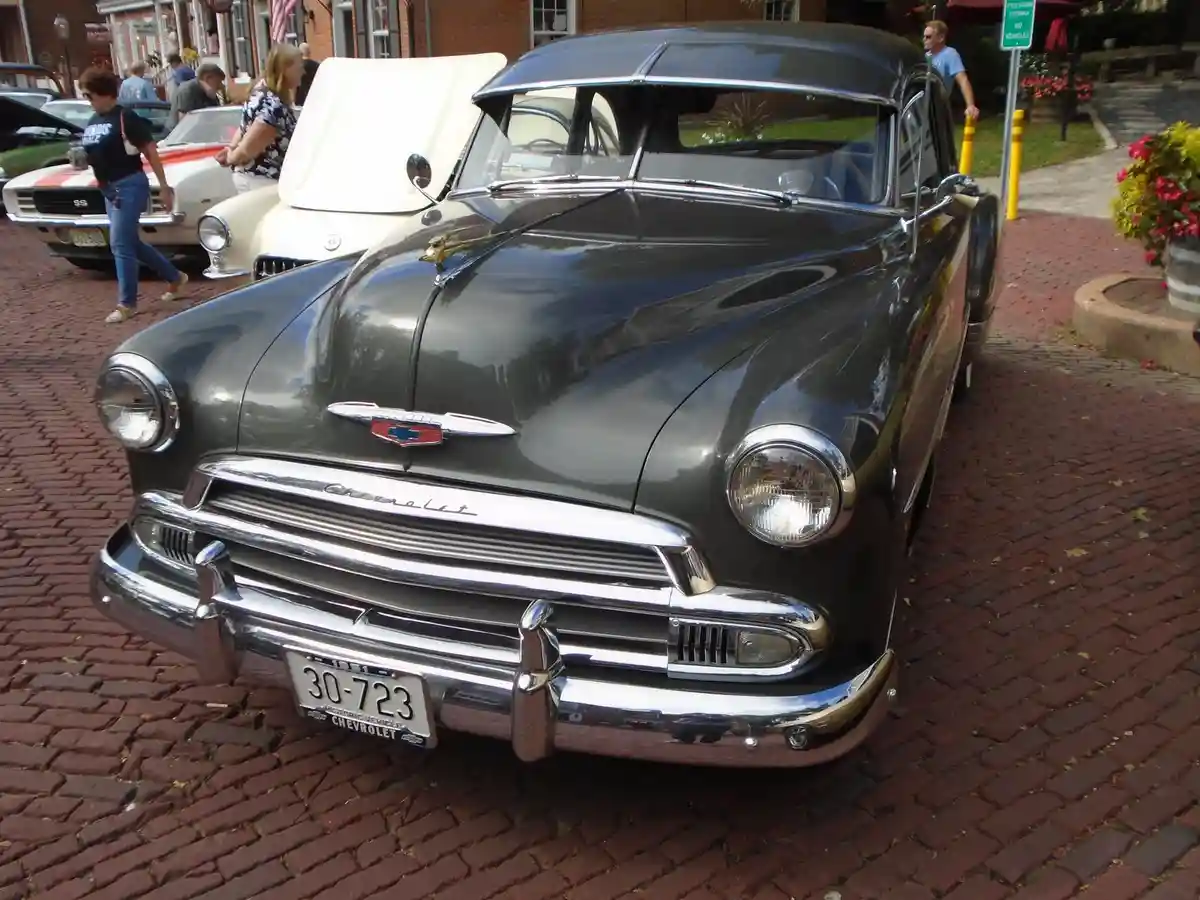 A shiny black 1951 Chevrolet is parked on a red brick street at a car show, with its chrome grille and bumper gleaming…