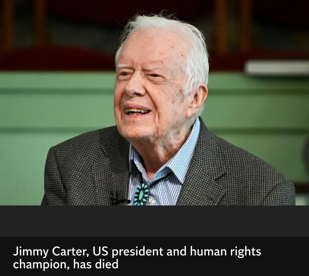 An elderly white man with white hair, wearing a dark checkered suit jacket over a blue and white striped shirt with a…