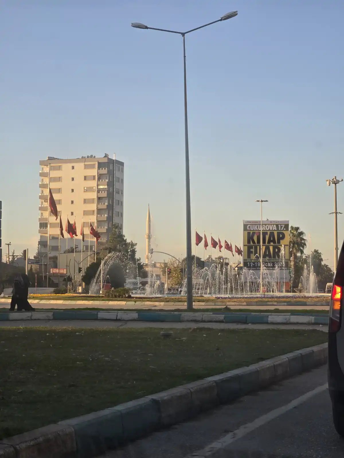 A daytime cityscape photo shows a roundabout with fountains, Turkish flags, a tall building, and a mosque minaret in the…