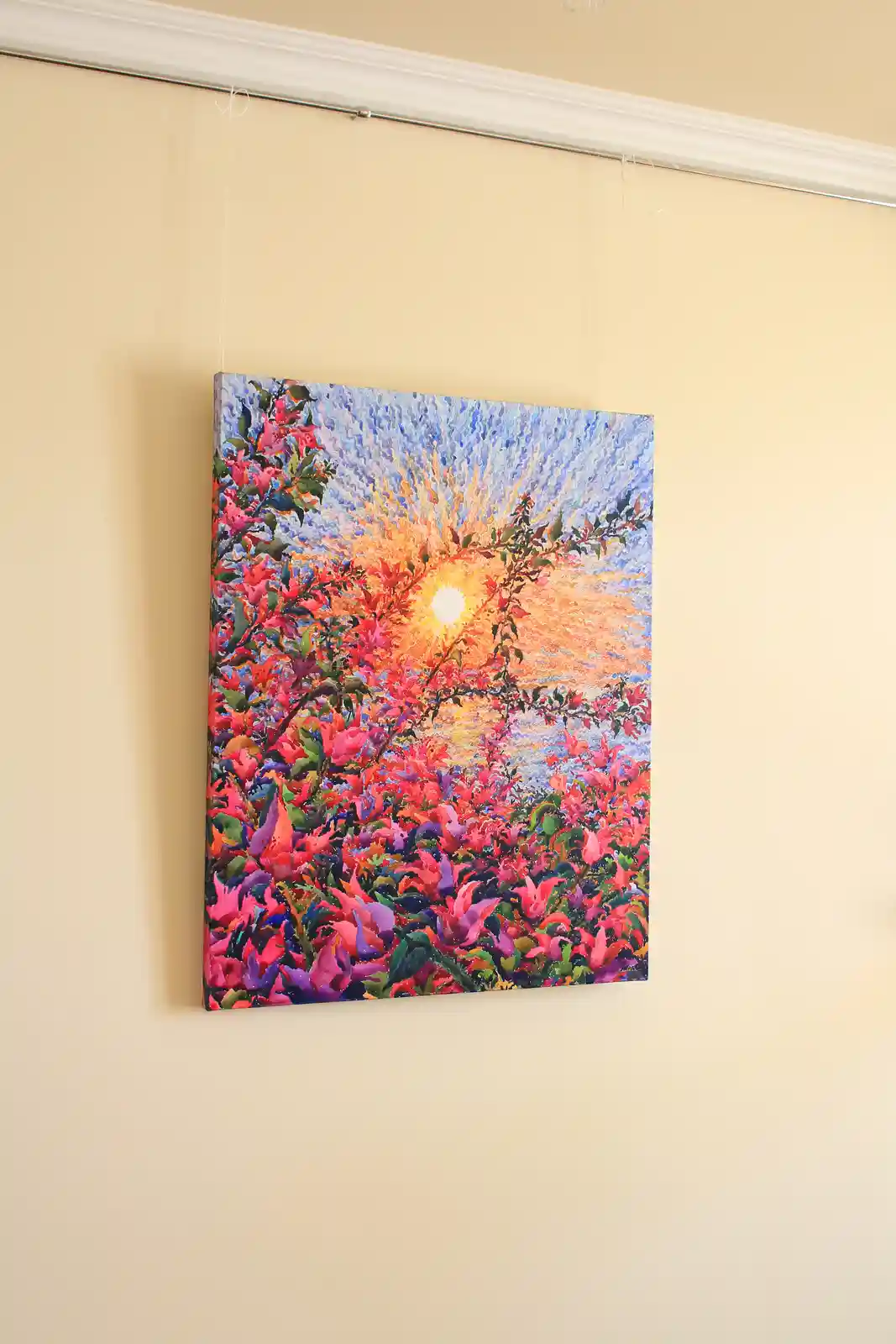 A vibrant, colorful painting of a sunset with pink and purple flowers and a bright sun is hanging on a beige wall by thin…