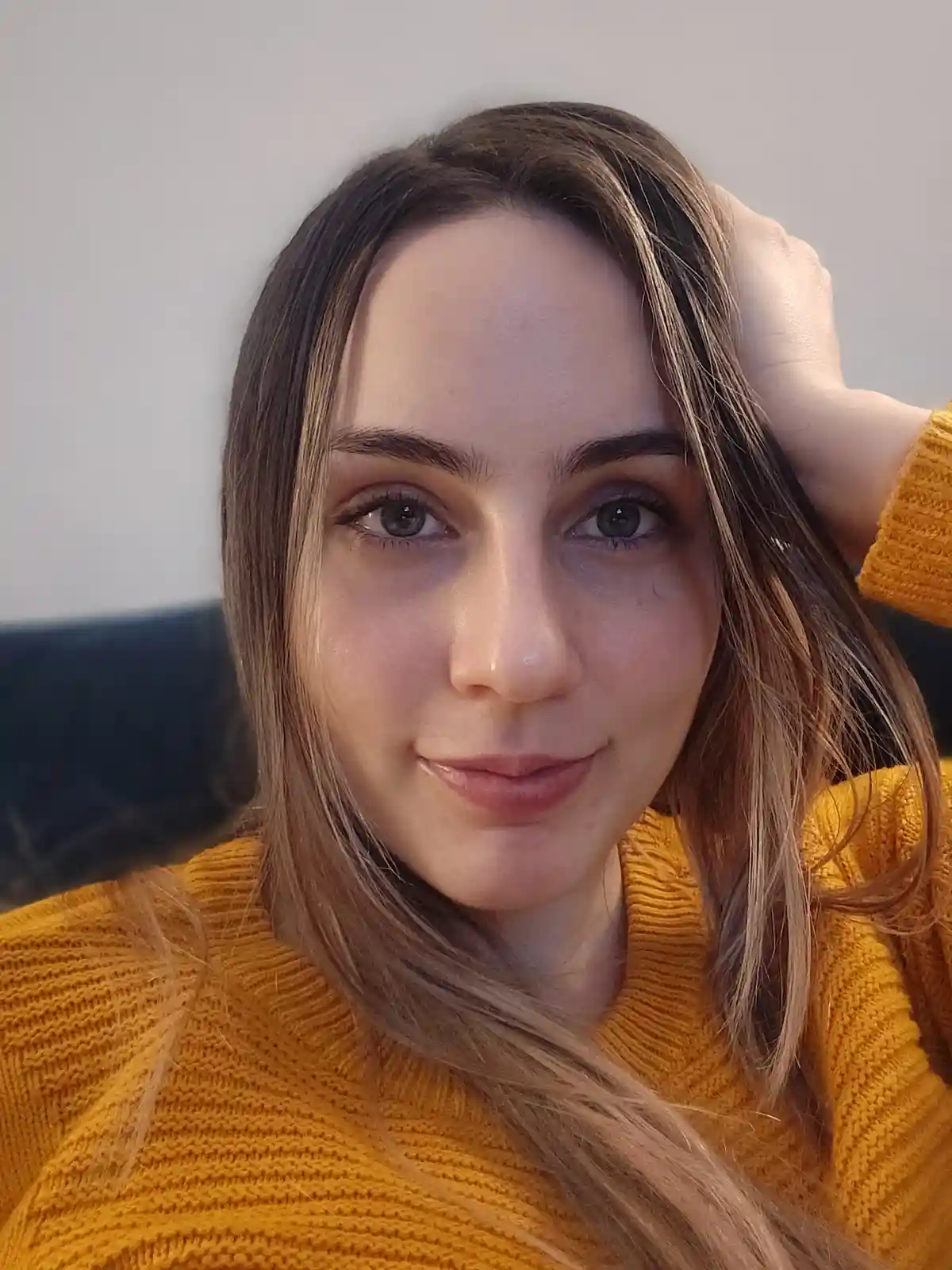 A woman with long brown hair and a yellow sweater looks at the camera with a slight smile while her hand is behind her head…