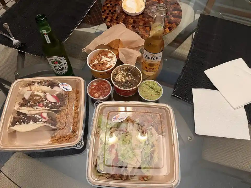 A table is covered with two large takeout containers of tacos, side dishes including salsa, guacamole, beans, and rice, and…