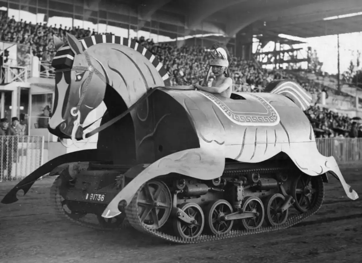 A black-and-white historical photograph shows a man in a helmet seated atop a military tank that has been elaborately…