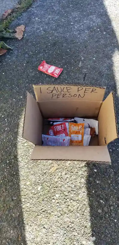 A cardboard box sits open on a gravel path, filled with various ketchup and hot sauce packets, including red 'FIRE!' and…