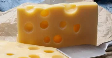 A close-up photo of a large, yellow block of Swiss cheese with characteristic holes, resting on crumpled brown paper. A…