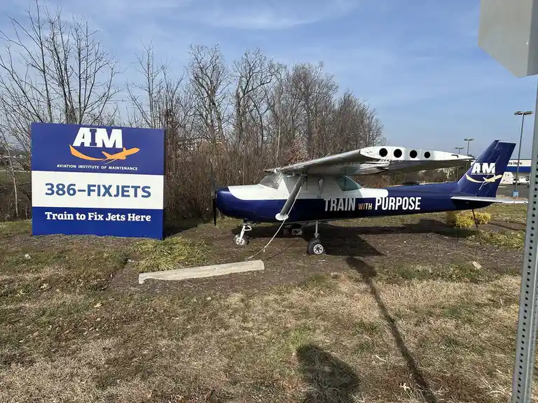 A small blue and white airplane sits parked next to a large blue sign for the Aviation Institute of Maintenance. The sign…