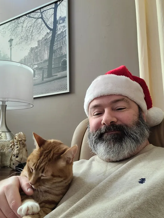 a man with a gray beard wearing a red and white santa hat sits in a chair holding a sleeping orange tabby cat. He has his…