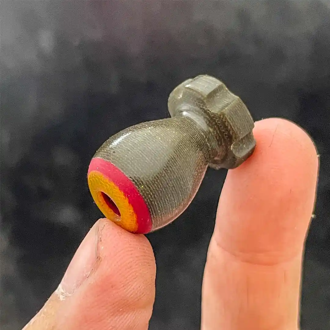 a close-up photo shows a person holding a small, gray 3D-printed airsoft BB between their thumb and index finger. The BB has…