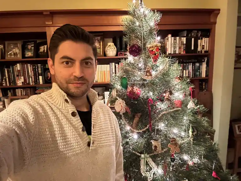 A man in a cream-colored knit sweater stands in front of a decorated Christmas tree with various ornaments, including a…