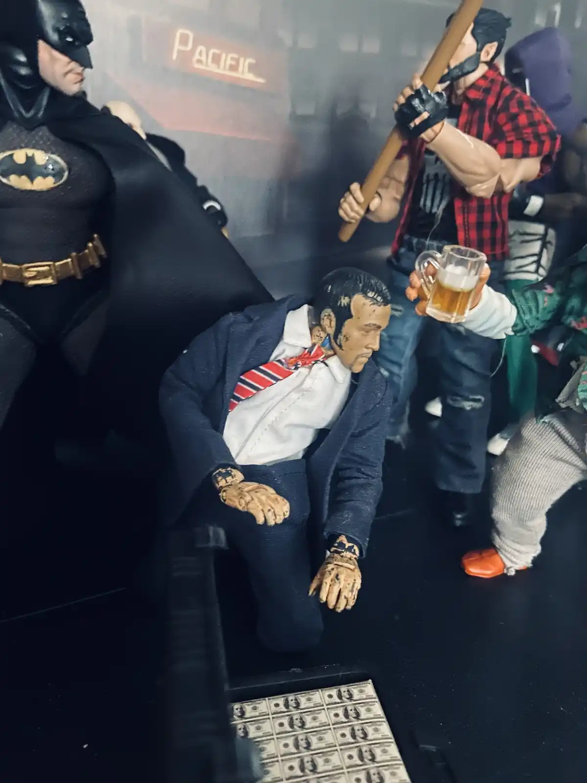 A photo of action figures: Batman stands menacingly, while a man in a suit kneels on the ground holding a beer mug…