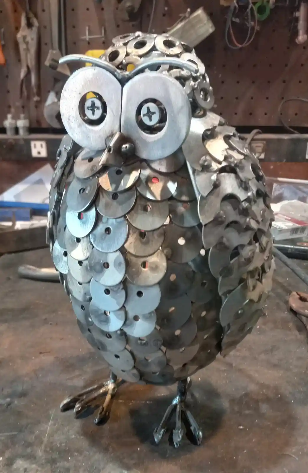 A handmade metal owl sculpture sits on a workbench, constructed from numerous small, circular metal discs welded together to…