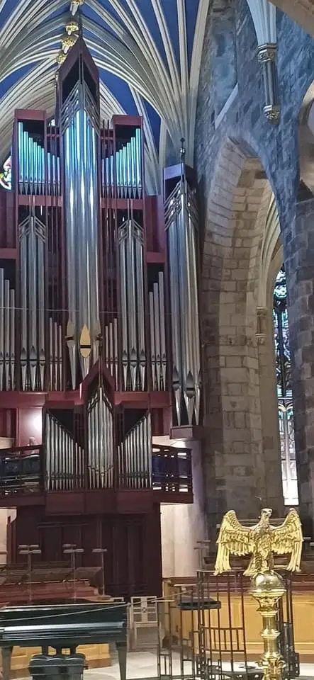 A large, ornate pipe organ with red wooden casework and silver pipes stands inside a stone church with arched ceilings and…