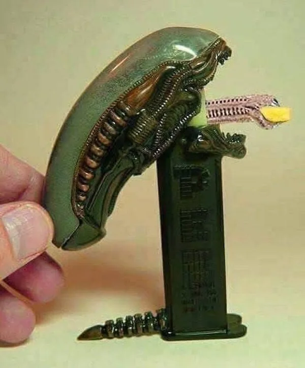 A hand holds a small green alien head toy, which is mounted on top of a black magazine. The alien's mouth is open, revealing…