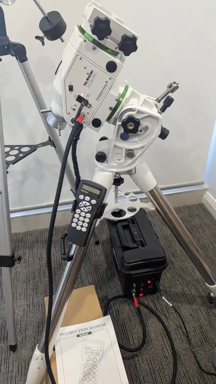 A white Sky-Watcher AZEQ6 PRO telescope mounted on a tripod indoors, with its SynScan hand controller attached and a printed…