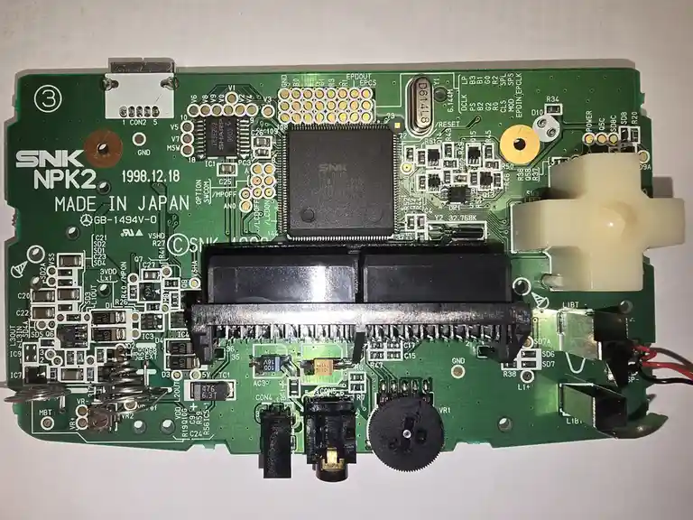 A top-down photo of a green SNK NPK2 arcade game circuit board from December 18, 1998, made in Japan, showing various…