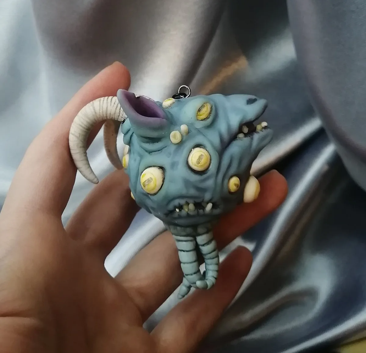 A hand holds a small, grotesque blue figurine with multiple yellow eyes, horns, and a toothy grin, resembling a horror…