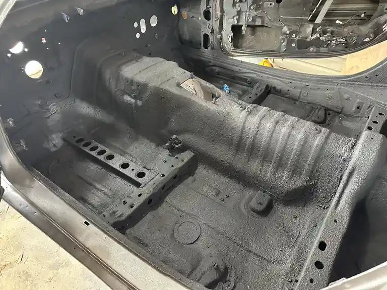 The image shows the interior of a car's trunk or cargo area, stripped down to bare metal with a rough, textured black…