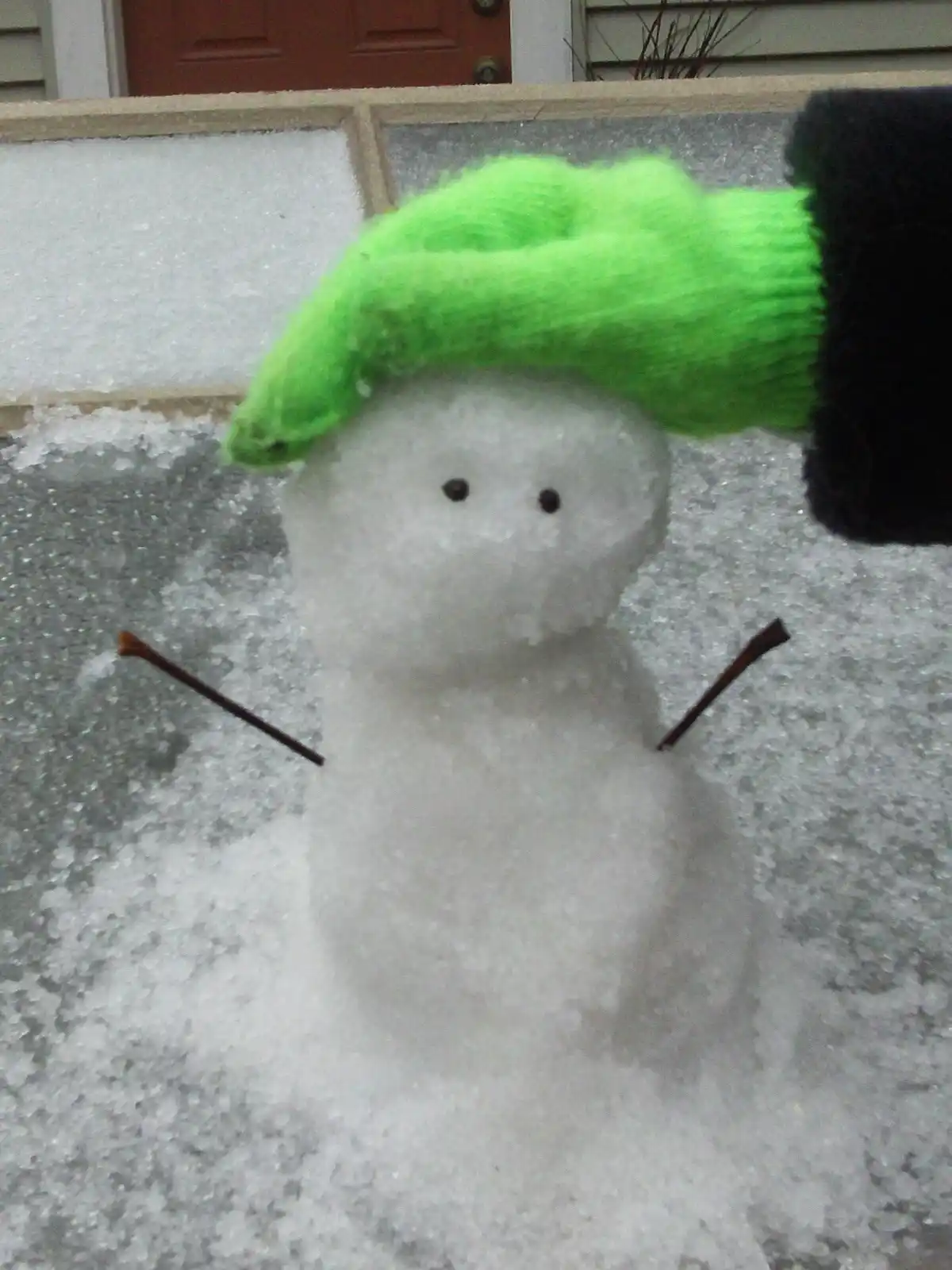 A small snowman made of two snowballs with twig arms and black button eyes stands in snow, with a bright green knitted hat…