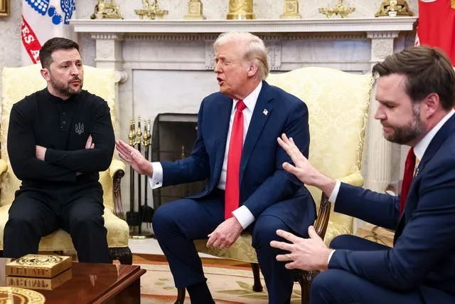 Three men sit in ornate chairs in a formal room with a fireplace and flags; the man in the center gestures while speaking…