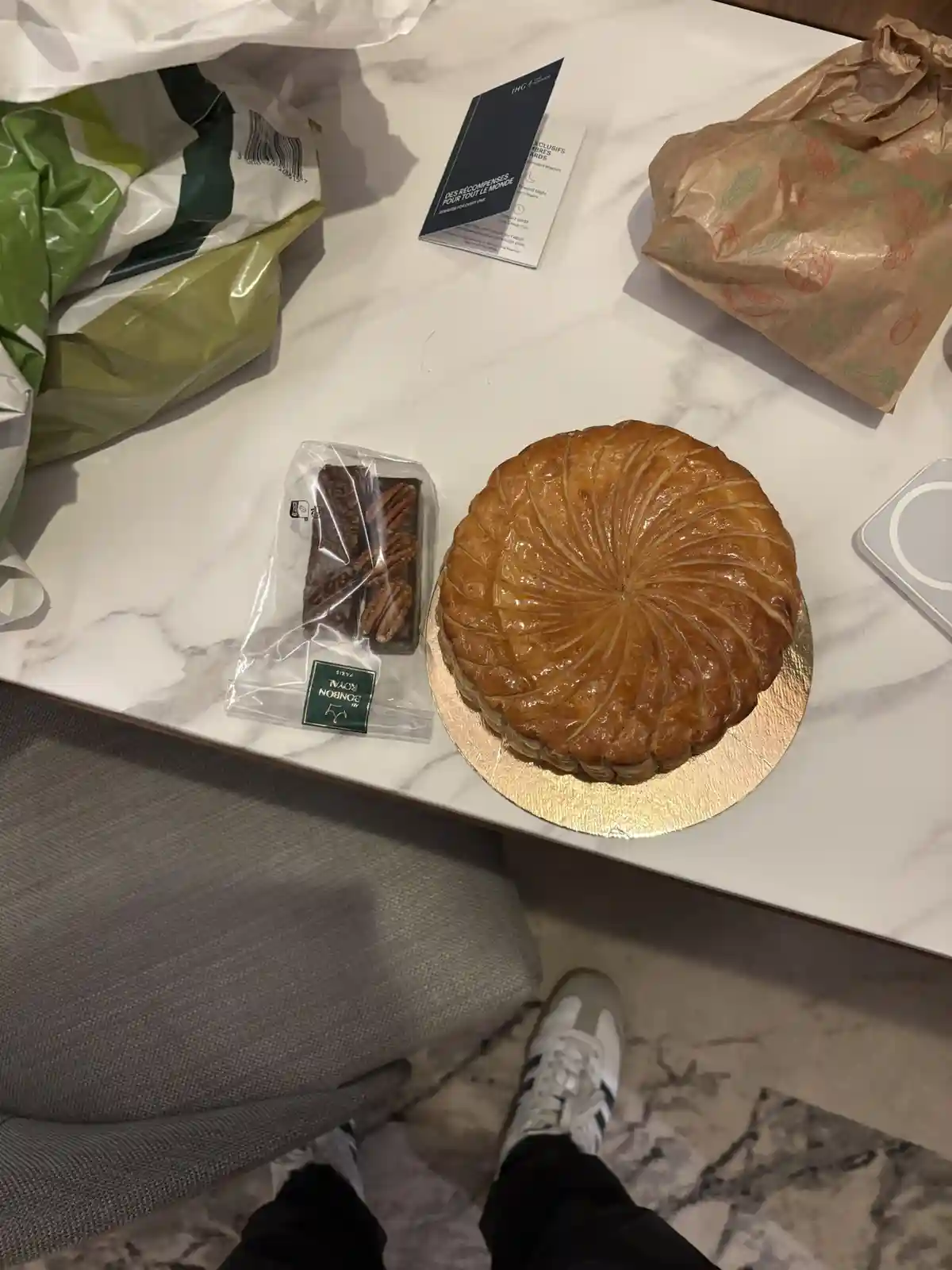 A top-down photo shows a marble countertop with a large, golden-brown, flaky pastry on a gold plate, next to a small package…