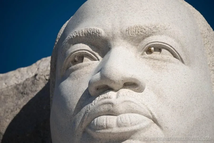 A close-up photograph of a large stone statue of Martin Luther King Jr., showing his face with a serene expression against a…