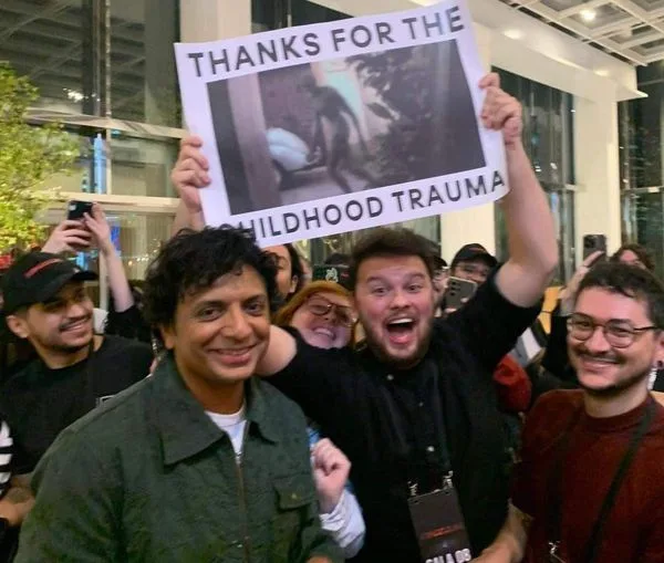 A man in a black shirt holds up a sign that reads 'THANKS FOR THE CHILDHOOD TRAUMA' with a photo of a monkey on it. He is…