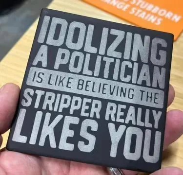 A hand holds a black square magnet with silver text that reads 'IDOLIZING A POLITICIAN IS LIKE BELIEVING THE STRIPPER REALLY…