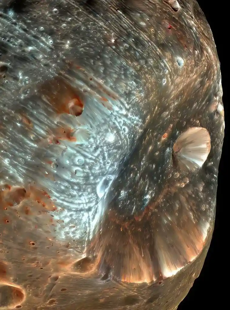 This image shows a close-up of the surface of a moon, likely Europa, featuring a large impact crater with a fan-shaped…