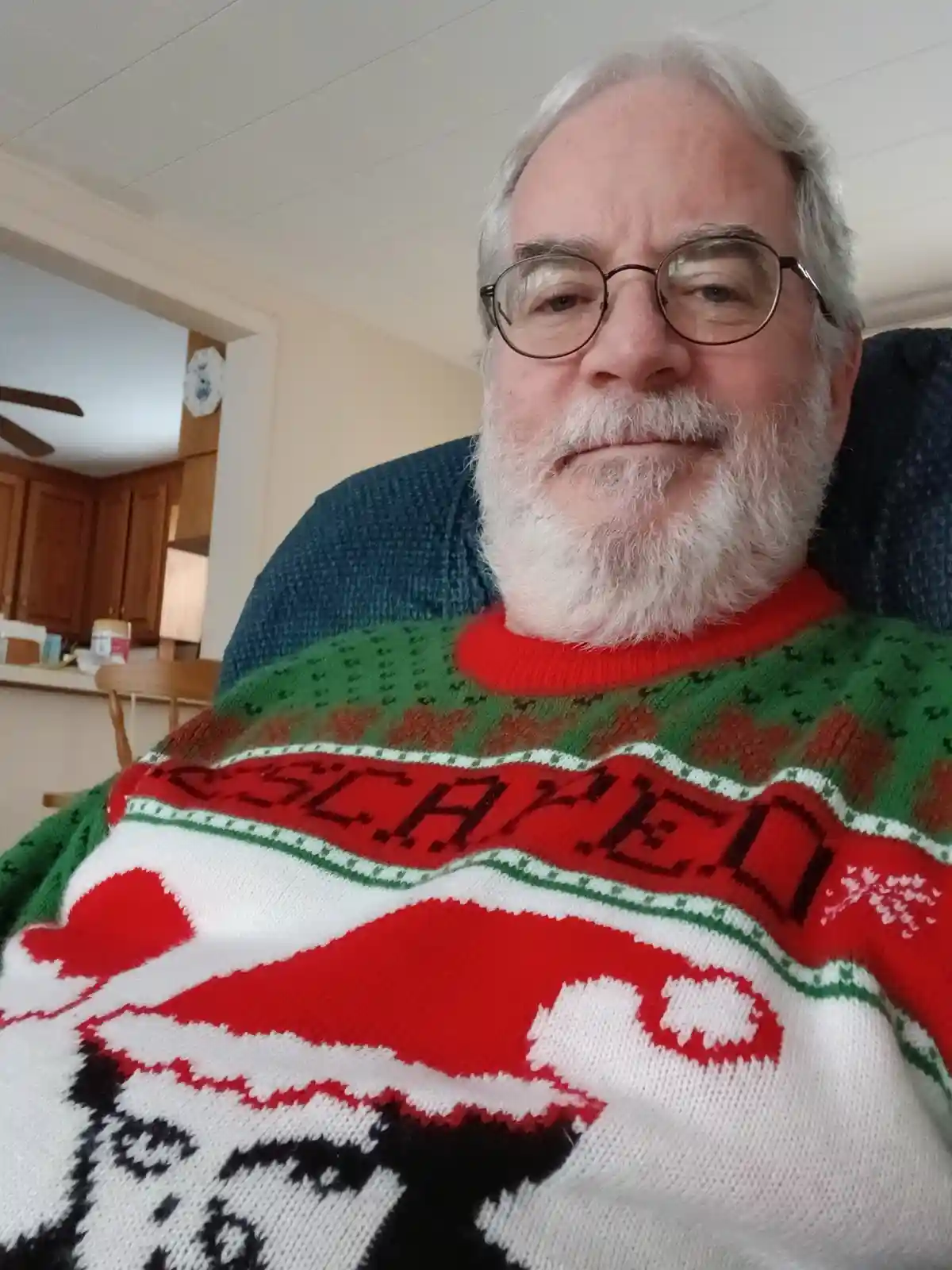 A close-up selfie of an older man with white hair, glasses, and a full white beard, wearing a festive red, green, and white…