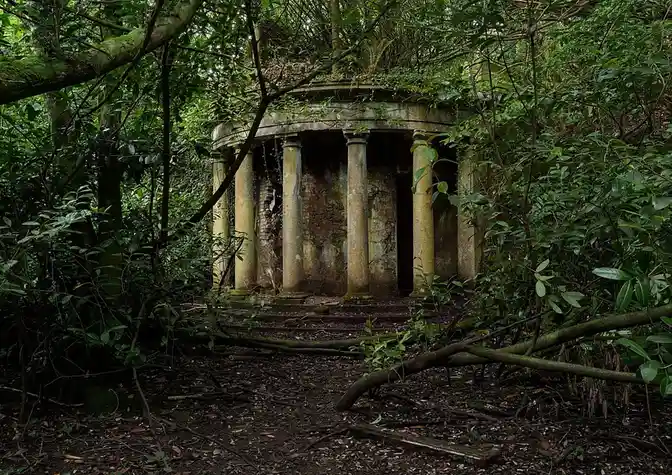 An overgrown, dilapidated stone structure with columns, partially collapsed and surrounded by dense green foliage and fallen…