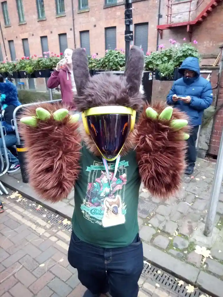 A person in a full-body furry costume with large brown furry arms ending in green claws, wearing a yellow-framed visor and a…
