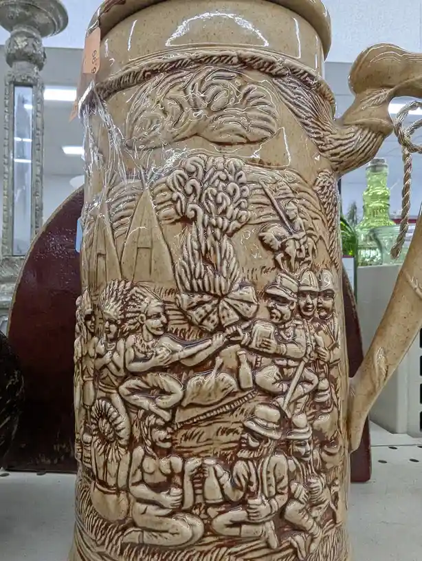 a close-up of a ceramic beer stein wrapped in plastic, featuring a detailed relief scene with indigenous people and…