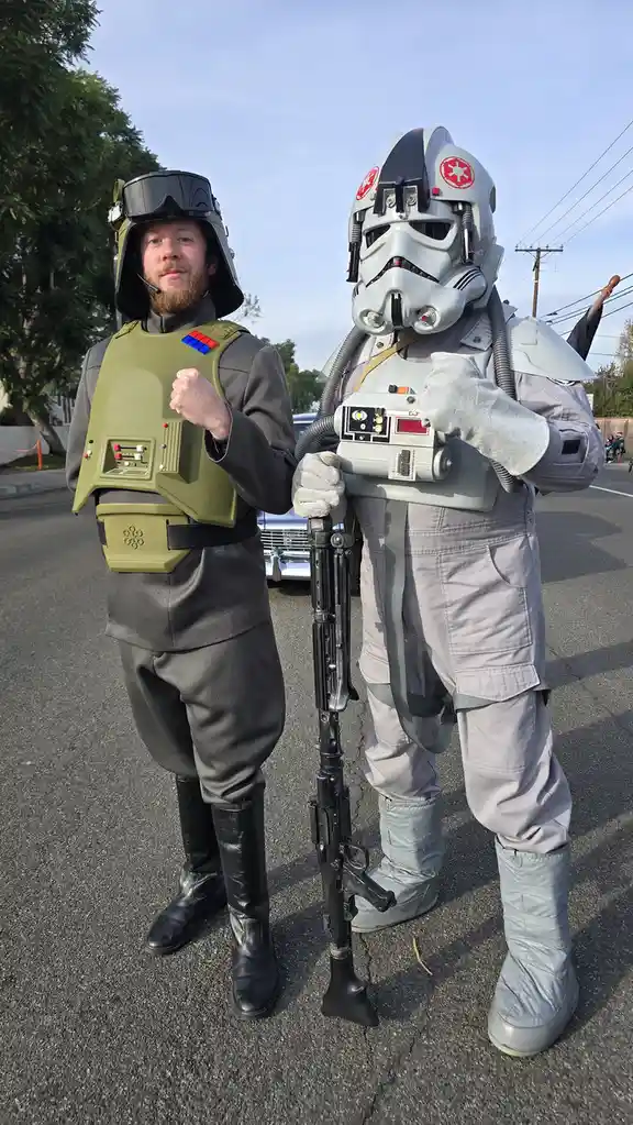 Two men in Star Wars costumes stand side-by-side on a street. The man on the left wears a green vest over grey clothes, a…