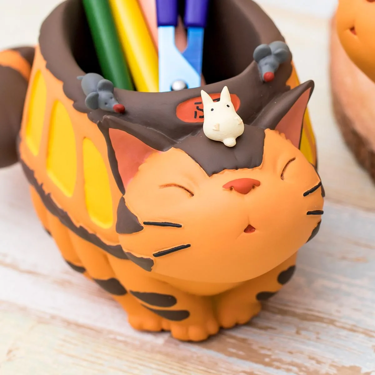 a ceramic pencil holder shaped like a sleeping orange cat with black stripes, featuring a small white creature and two gray…