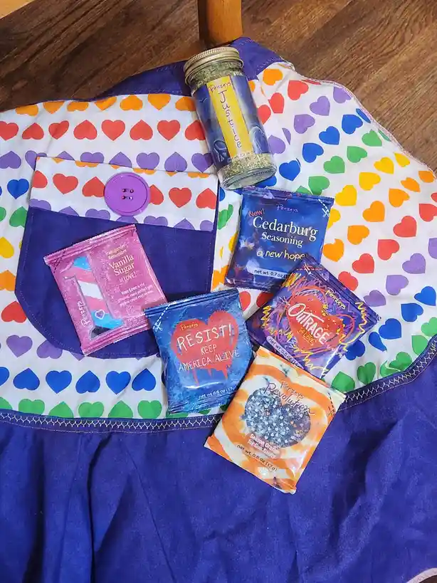 A colorful heart-patterned apron holds several small bags and a jar of Penzeys spice blends, each with satirical political…