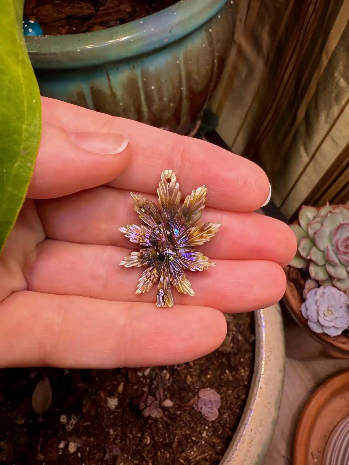 A hand holds a small, iridescent, leaf-shaped object resembling a cannabis leaf, with shimmering gold and purple hues. The…