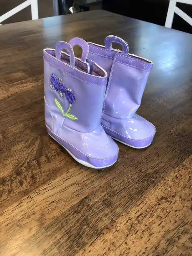 A pair of shiny, light purple toddler rain boots with floral appliqués sits on a dark wooden table. The left boot features a…
