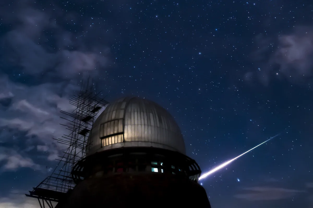 A nighttime photo shows a domed observatory with scaffolding on its side, set against a starry sky with clouds. A bright…