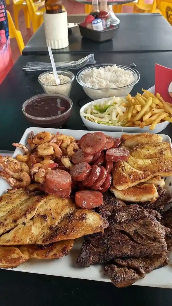 A large white plate piled high with various grilled meats including chicken, steak, sausage, and shrimp, accompanied by side…
