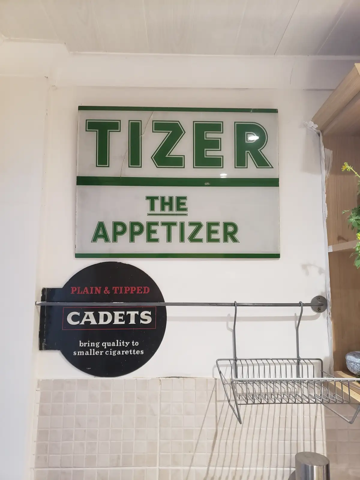 A vintage-style sign reads 'TIZER THE APPETIZER' in green letters on a white background, mounted above a black circular sign…